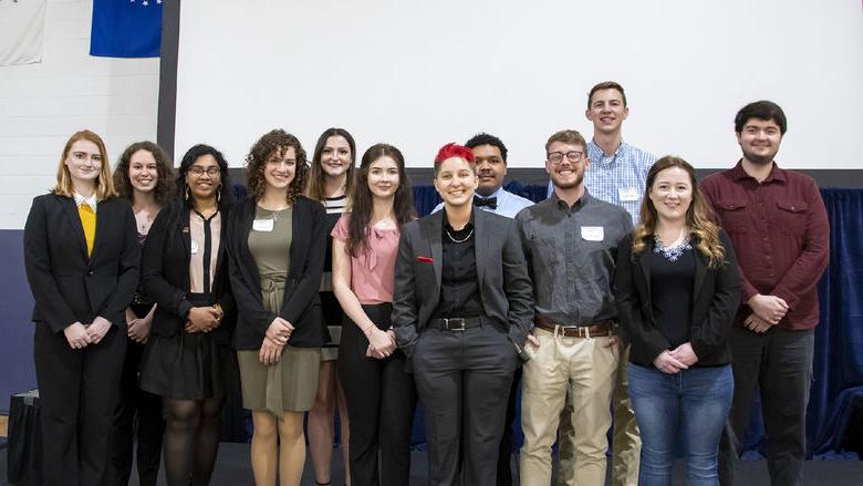 A dozen Penn State students pose for a photo on stage in front of a projector screen